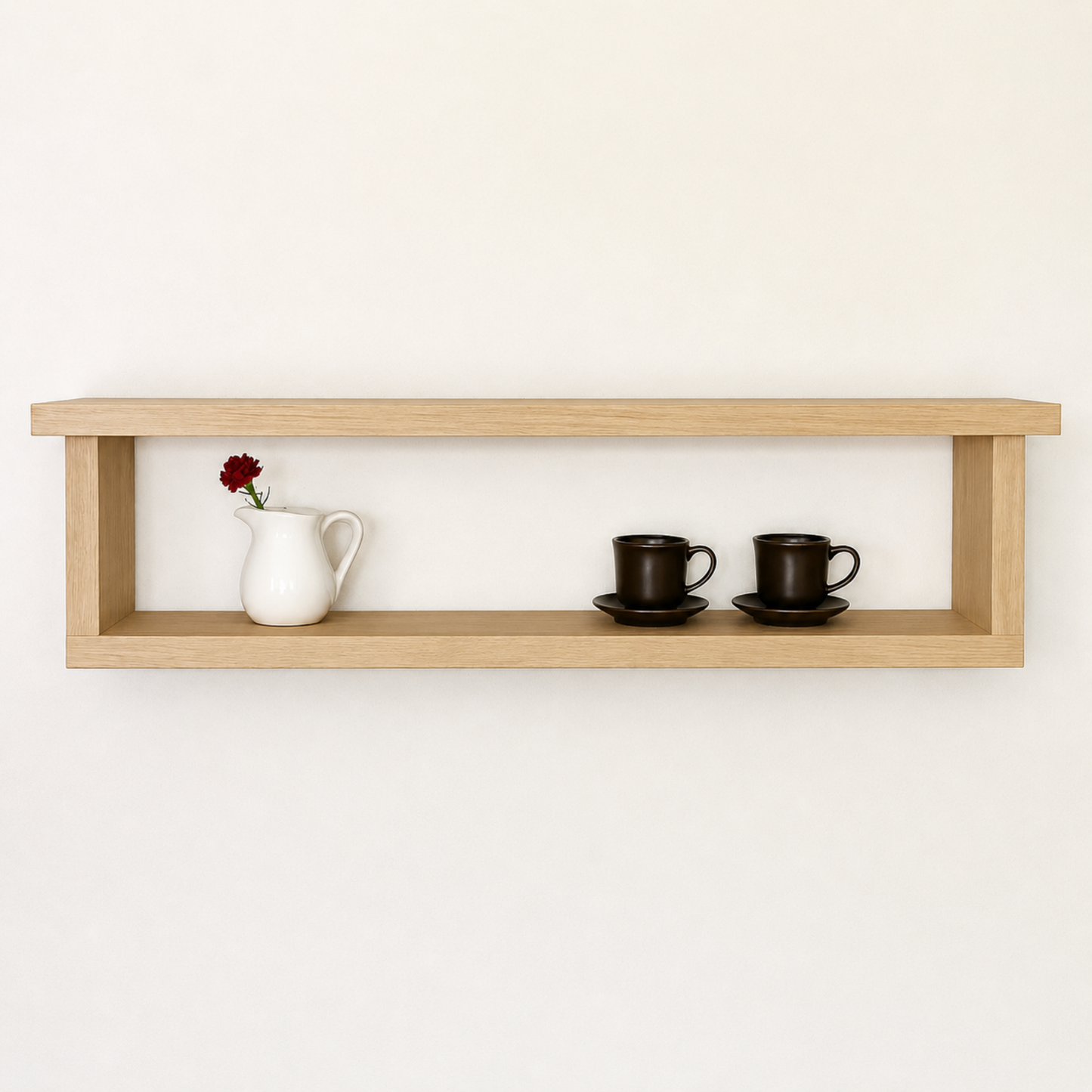 Wooden shelf with a white pitcher, two black cups on saucers, and a small red flower against a plain white background.