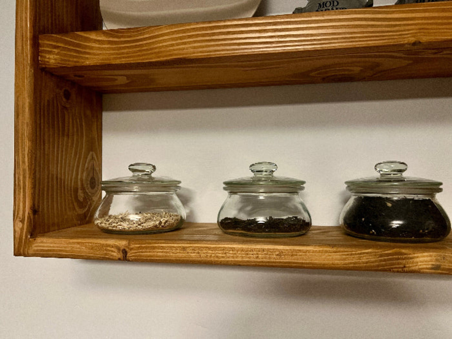 Three glass jars with lids on a wooden shelf against a white wall.