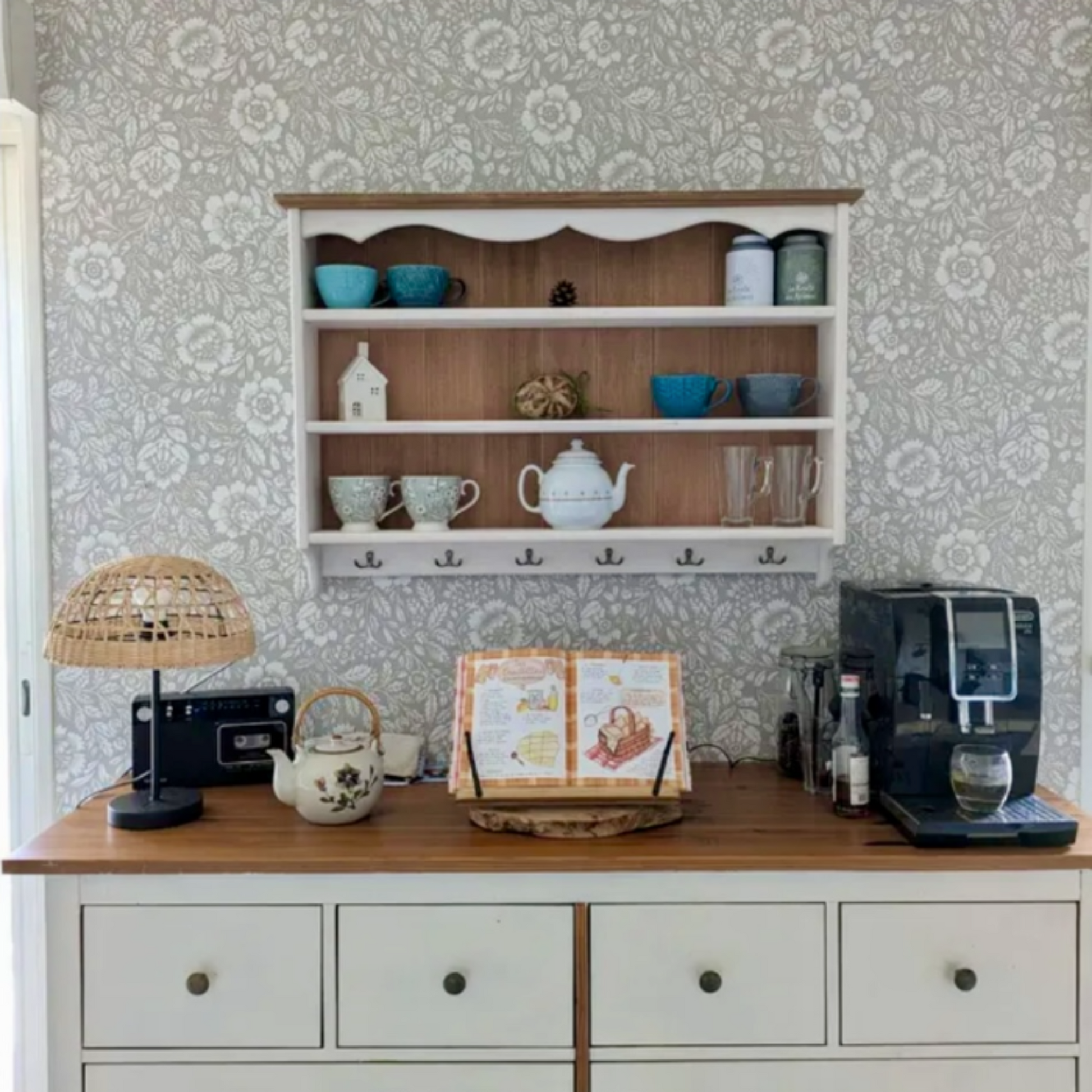 A handcrafted wooden shelf with a selection of mugs and teaware displayed on it, mounted on a wall with floral wallpaper.