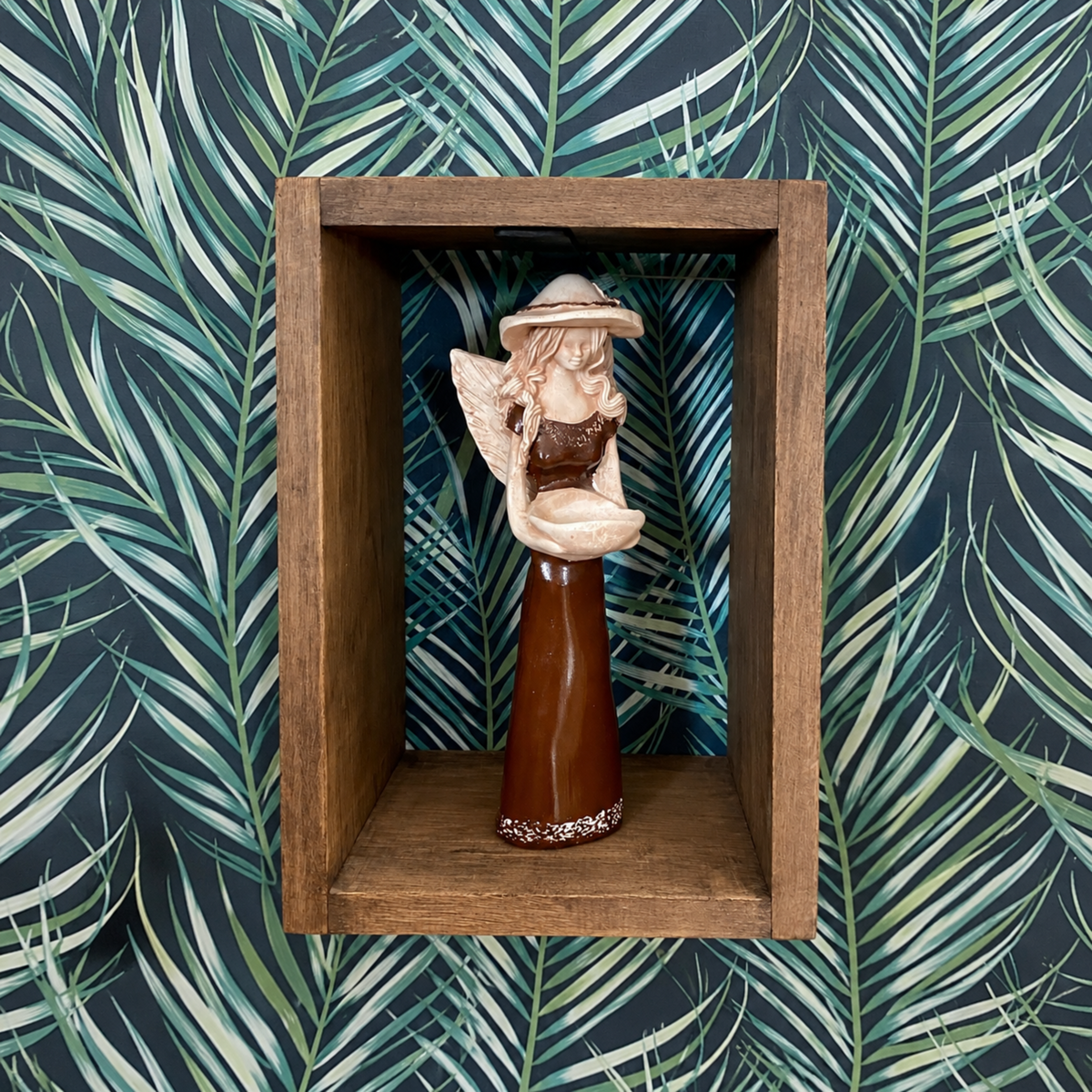 A small decorative angel figurine displayed inside a rectangular wooden shadow box, set against a dark tropical palm leaf patterned background.