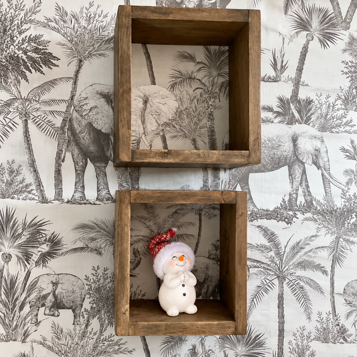 Two rustic wooden square wall boxes mounted on a tropical elephant-patterned wallpaper, with the bottom box containing a small white snowman figurine wearing a red hat.