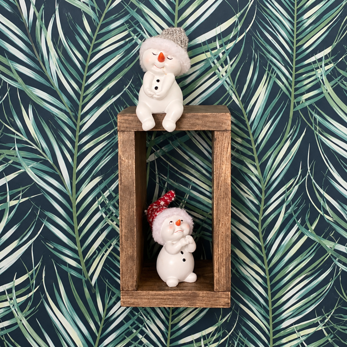 Two small white snowman figurines displayed on a rustic wooden shelf against a dark tropical palm leaf wallpaper background.