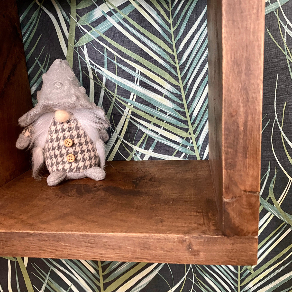 A small plush decorative gnome figurine with a gray hat and a brown-and-white houndstooth vest sits on a rustic wooden shelf against a black wallpaper background with a green palm leaf pattern.