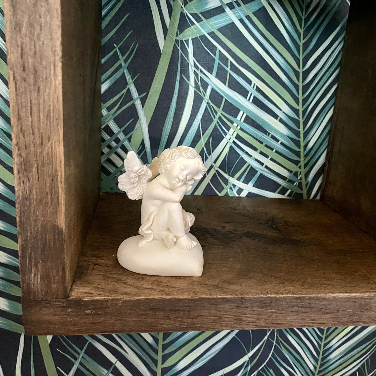 A small cream-colored cherub figurine sitting on a wooden shelf in front of a dark tropical palm leaf patterned backdrop.