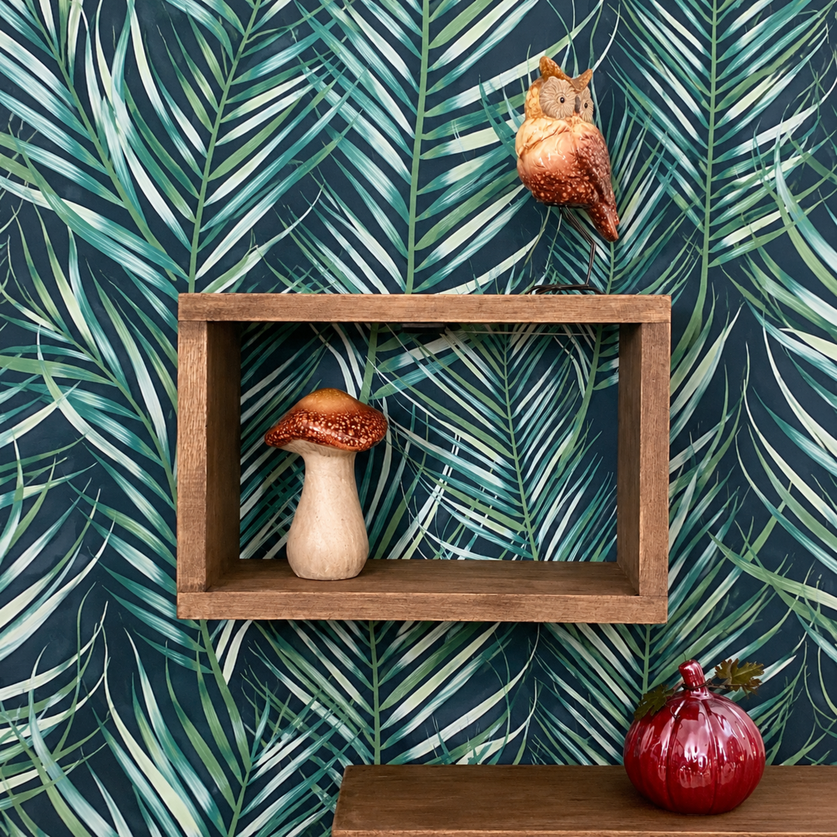 A styled interior scene featuring dark teal wallpaper with a green palm leaf pattern, a rustic wooden floating shelf, a ceramic mushroom, a hanging owl ornament, and a red decorative pumpkin on a wooden ledge.