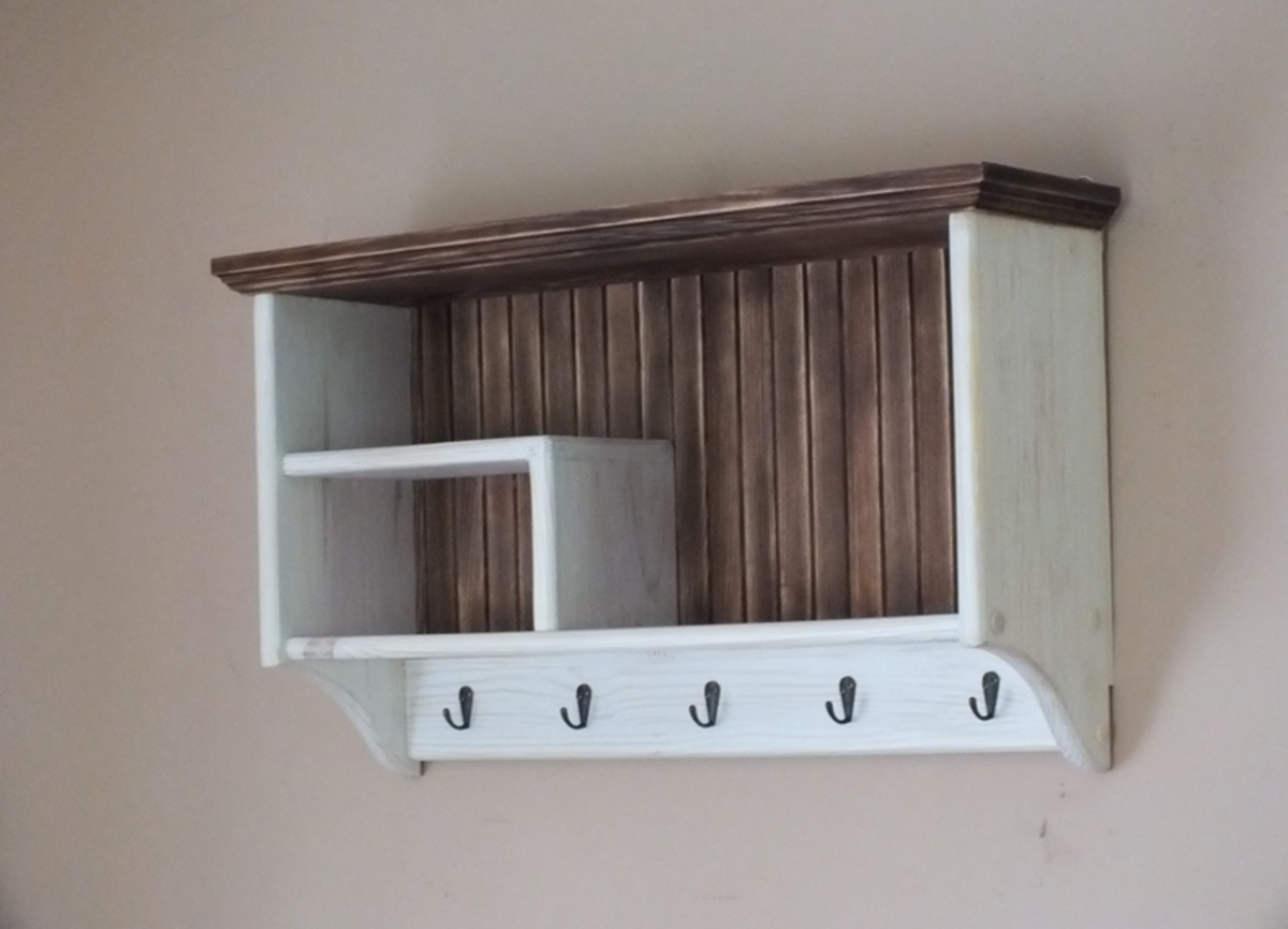 A rustic wall-mounted wooden storage shelf with a dark brown slatted back panel, a white open side compartment, a small white shelf, and five black hooks along the bottom, shown against a plain beige wall.