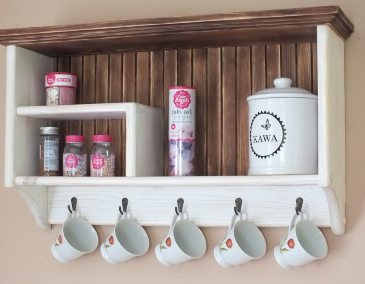A rustic white wooden wall-mounted shelf with a dark brown top frame, featuring a slatted wood backing. The shelf is styled with various kitchen items, including a white ceramic canister labeled 'KAWA', jars, and a cake mix canister, with five white ceramic mugs hanging from hooks along the bottom edge against a beige wall.