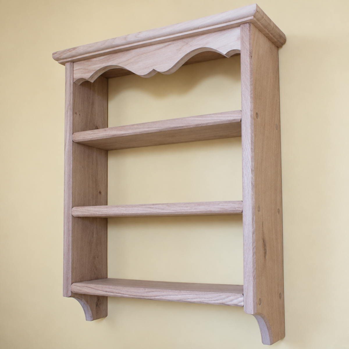A light natural wood wall-mounted bookshelf with three shelves, decorative molding on the top frame, and curved bottom brackets, shown against a pale yellow wall.