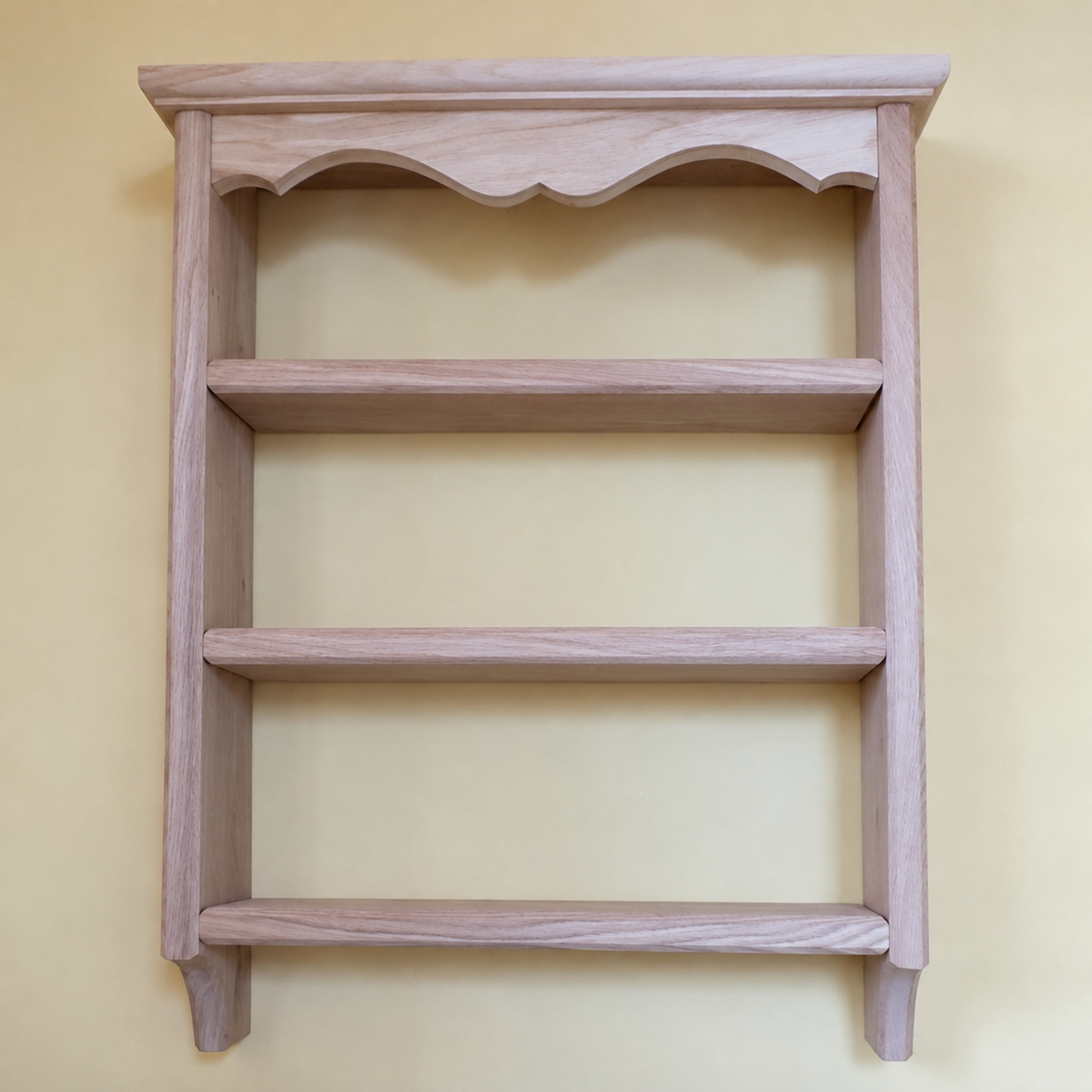A light natural wood wall-mounted shelf with three horizontal shelves and a decorative scalloped top frame, shown against a solid pale yellow background.