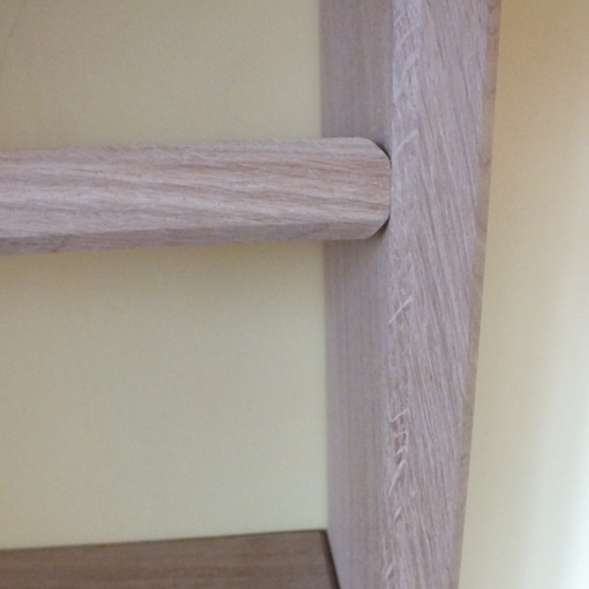 Close-up of a light oak wood shelf or ladder step showing the wood grain texture and joint against a pale yellow wall.