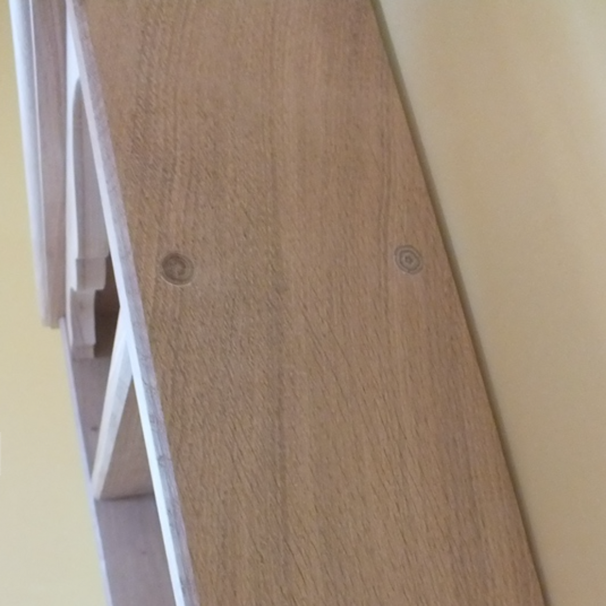 Close-up of a light oak wood shelf or ladder panel with visible screw heads, shown against a pale yellow wall.