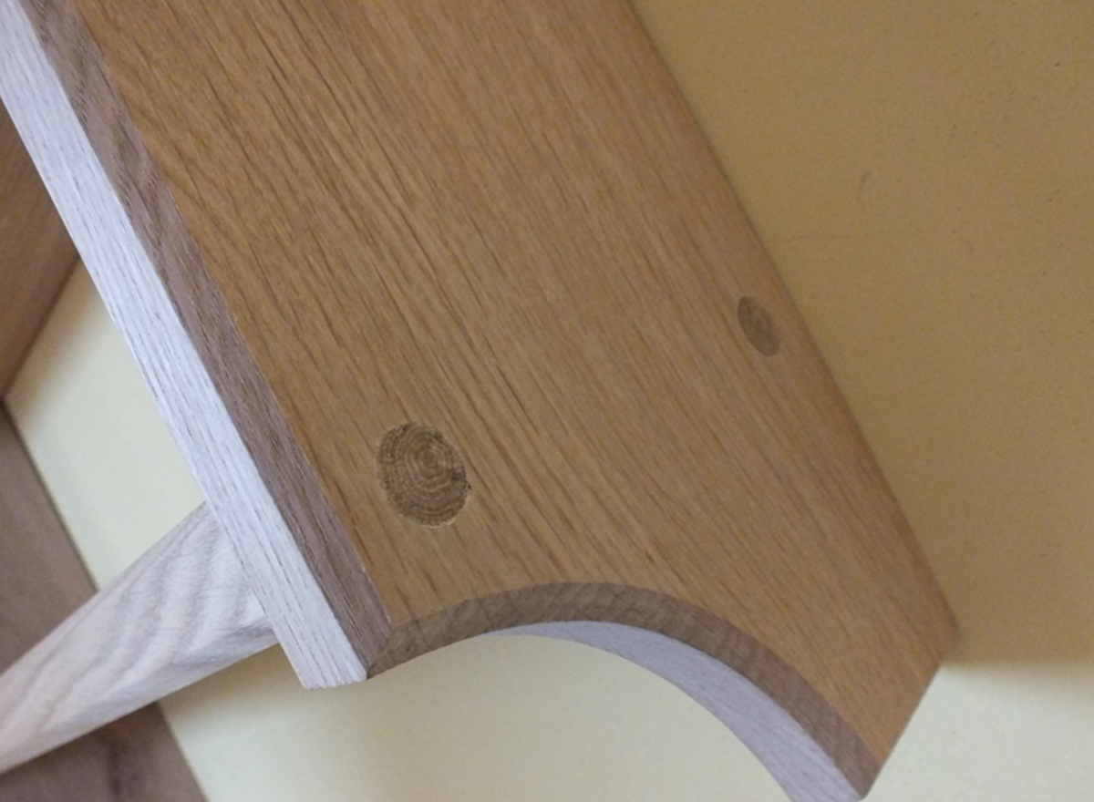 Close-up of a light oak wood chair or table leg with a curved apron and a visible wood grain knot, set against a pale cream background.