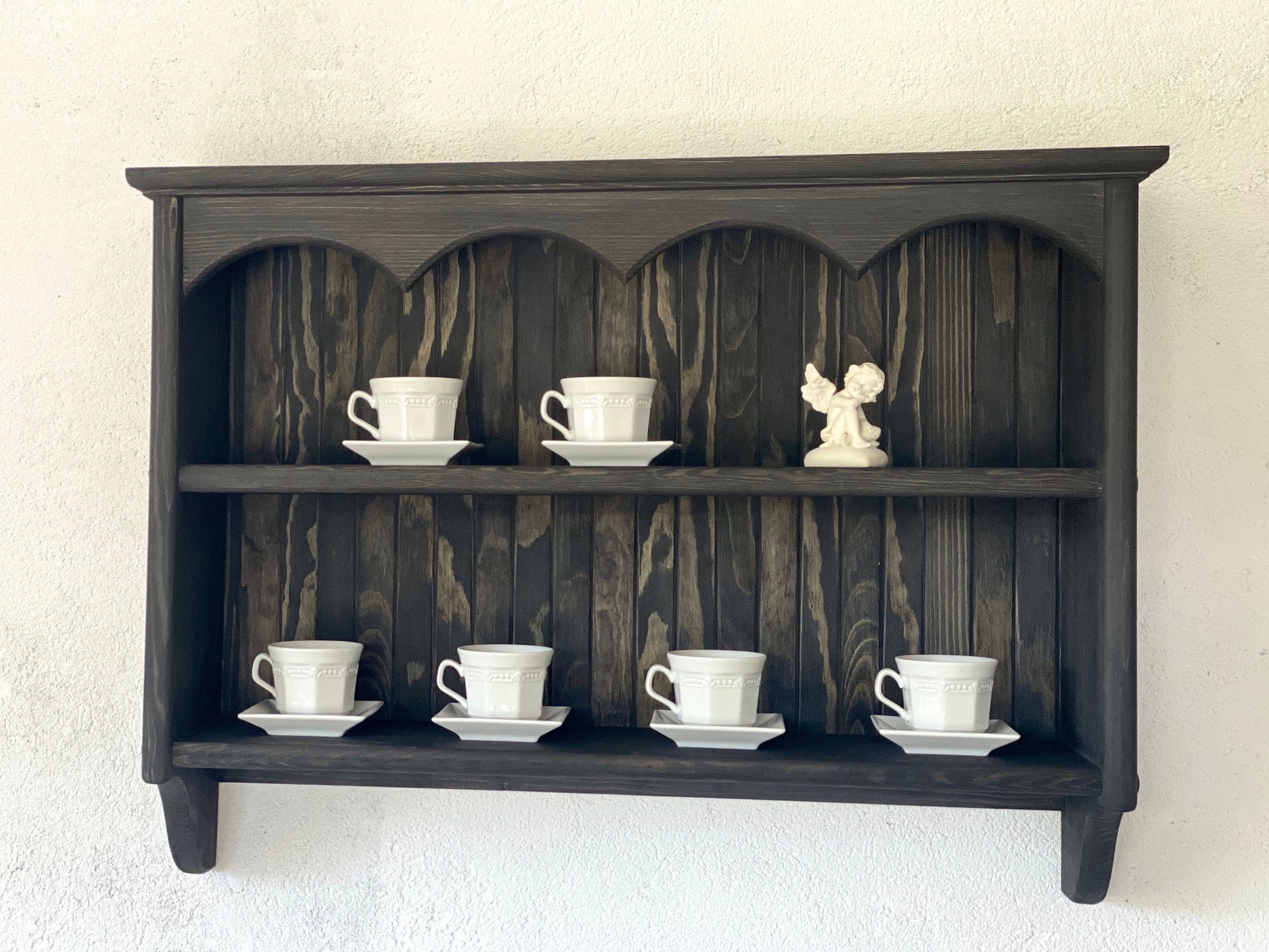 A wall mounted wooden shelving unit in a cottage style with a decorative top, displaying white cups on the shelves.