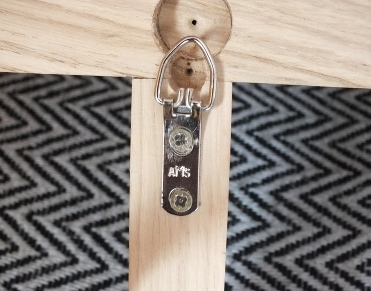 A close-up of a silver metal hanging hook or D-ring attachment mounted on a light wood board, with a black-and-white zigzag patterned surface in the background.