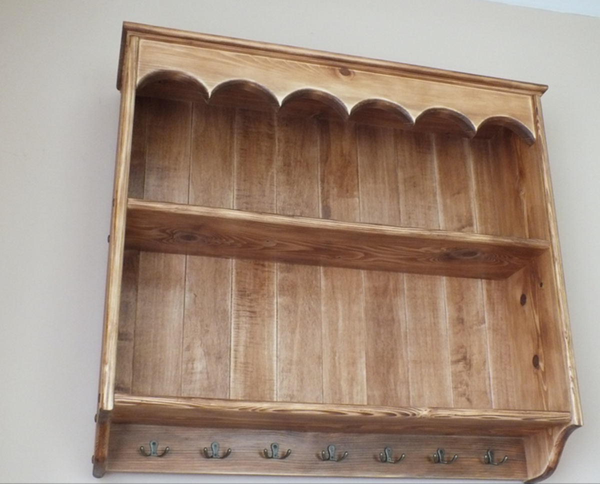 A rustic wooden wall-mounted shelf with a scalloped top edge, two interior shelves, and a row of small hooks along the bottom front, shown against a plain light wall.
