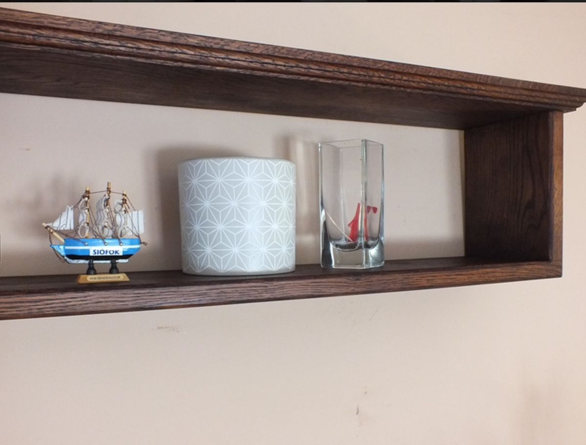A dark brown wooden open shelf mounted on a beige wall, styled with a small blue and gold model ship, a white patterned cylindrical container, and a clear glass vase containing a small red and white sailboat.