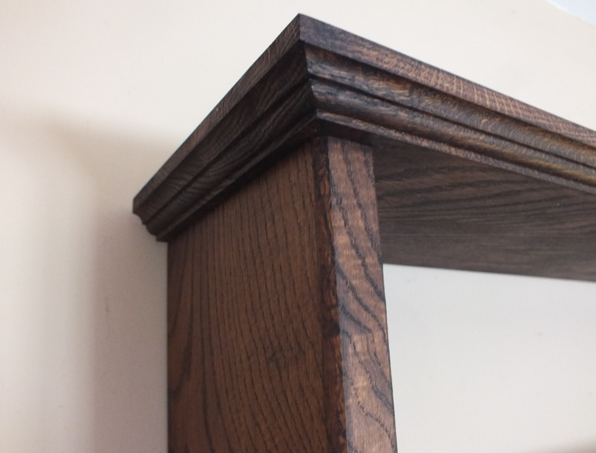 Close-up of a dark brown wooden shelf corner with a decorative molded top edge and visible wood grain, set against a plain off-white wall.