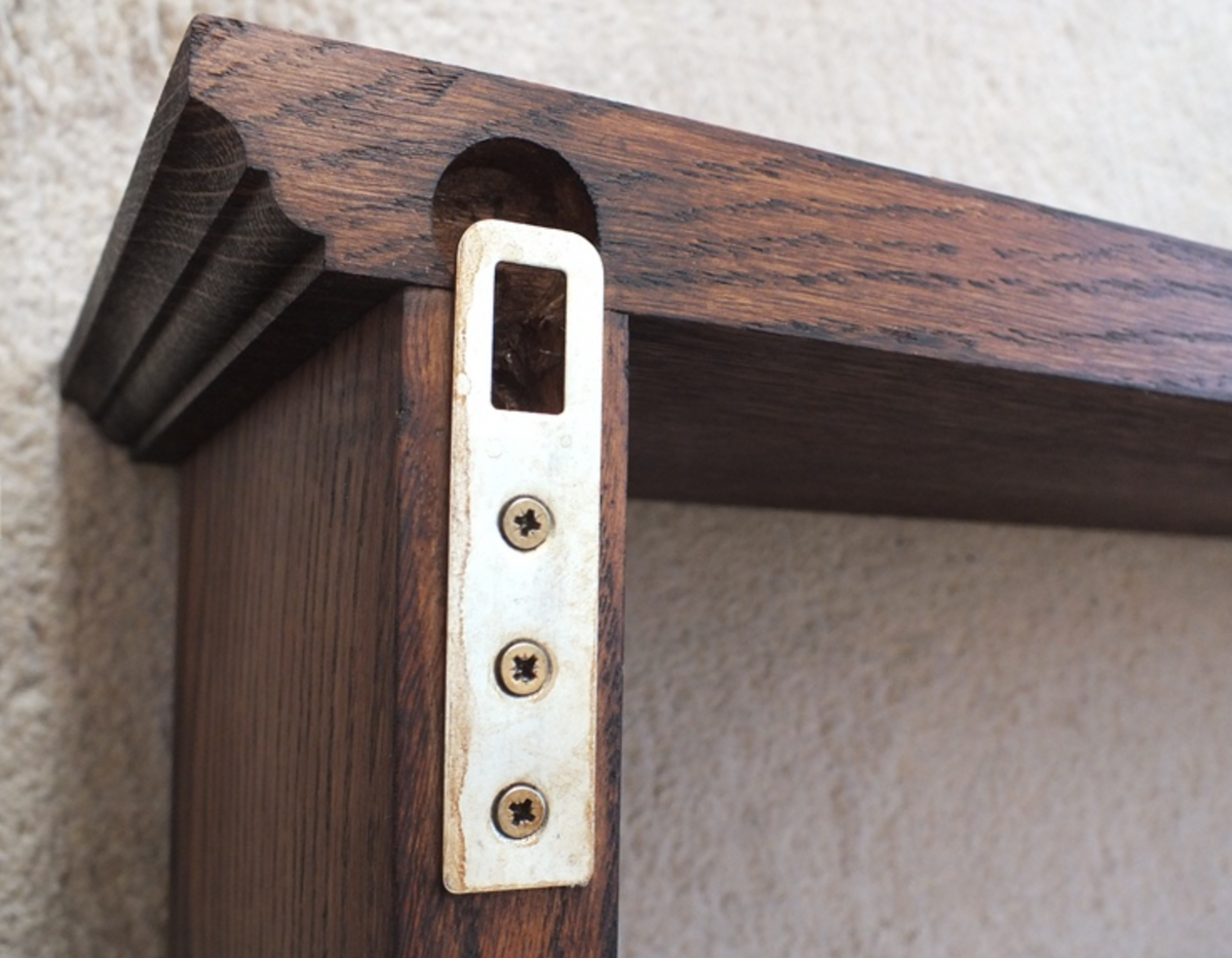 Close-up of a dark wood shelf corner with a brass metal bracket secured by three screws against a textured beige wall.