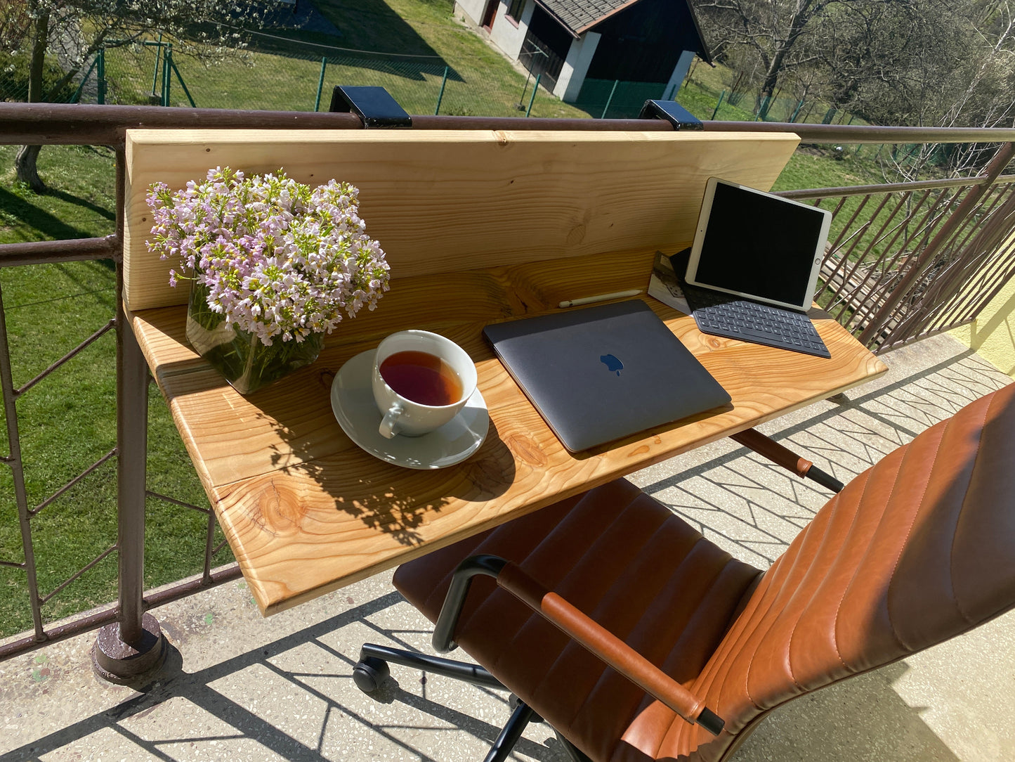Wooden outdoor desk with laptop, cup of tea, and flowers on a balcony.