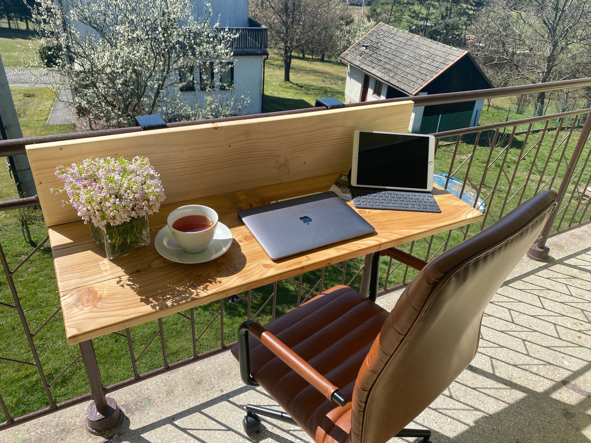 Wooden desk with laptop, cup of tea, and flowers on a balcony with a view of a house and trees.