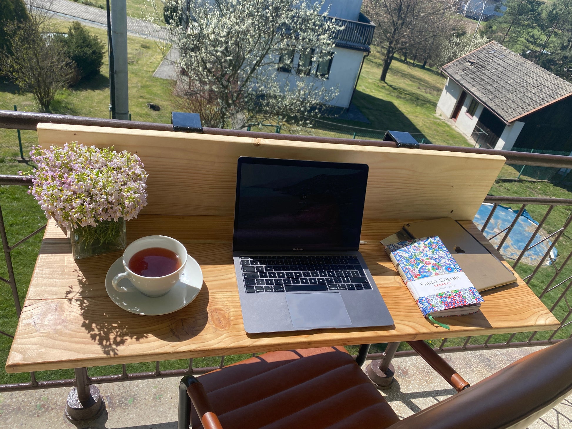Laptop on a wooden table with a cup of tea and flowers on a balcony