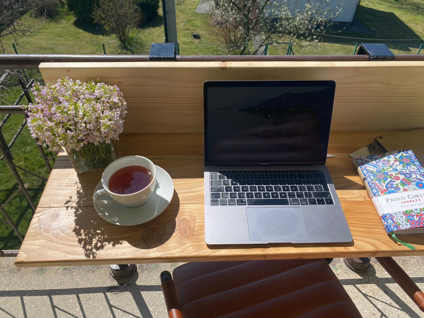 Laptop on a wooden table with a cup of tea and flowers on a balcony.
