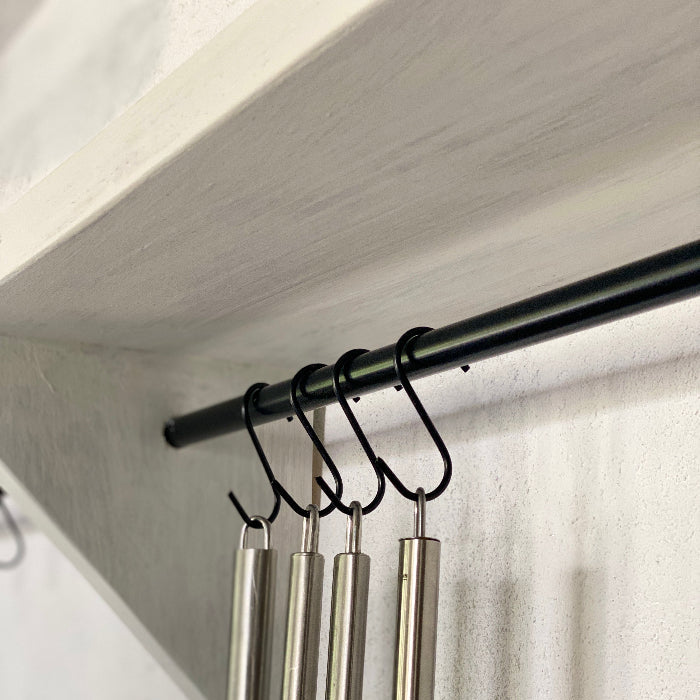 Metal whisks hanging from a black hook on a white shelf.