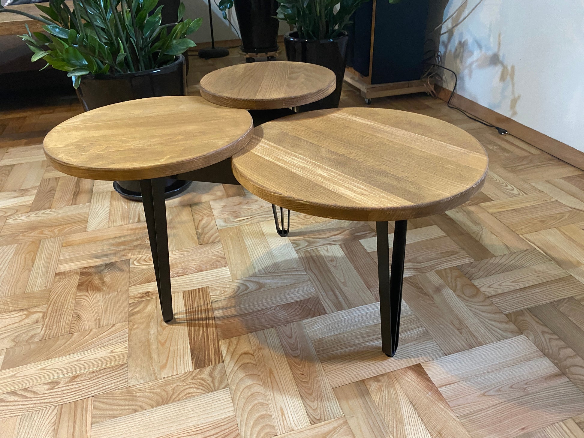 A round wooden coffee table with three tops and hairpin metal legs, displayed on a herringbone patterned floor.